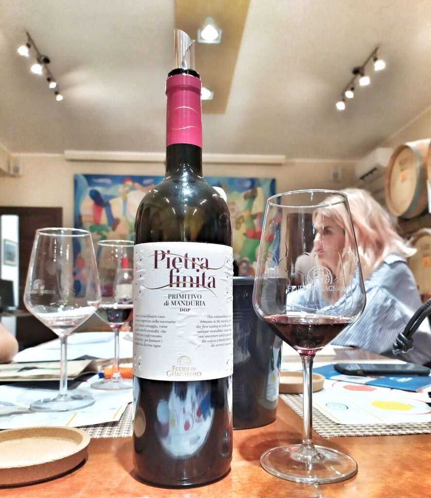 A professional indoor wine tasting scene at Feudi di Guagnano winery in Puglia. In the foreground, a bottle of "Pietra Finita" Primitivo di Manduria stands next to a half-filled glass of red wine. In the background, a man and a woman participate in a tasting session around a wooden table, with oak barrels (barriques) and a colorful modern painting on the walls.