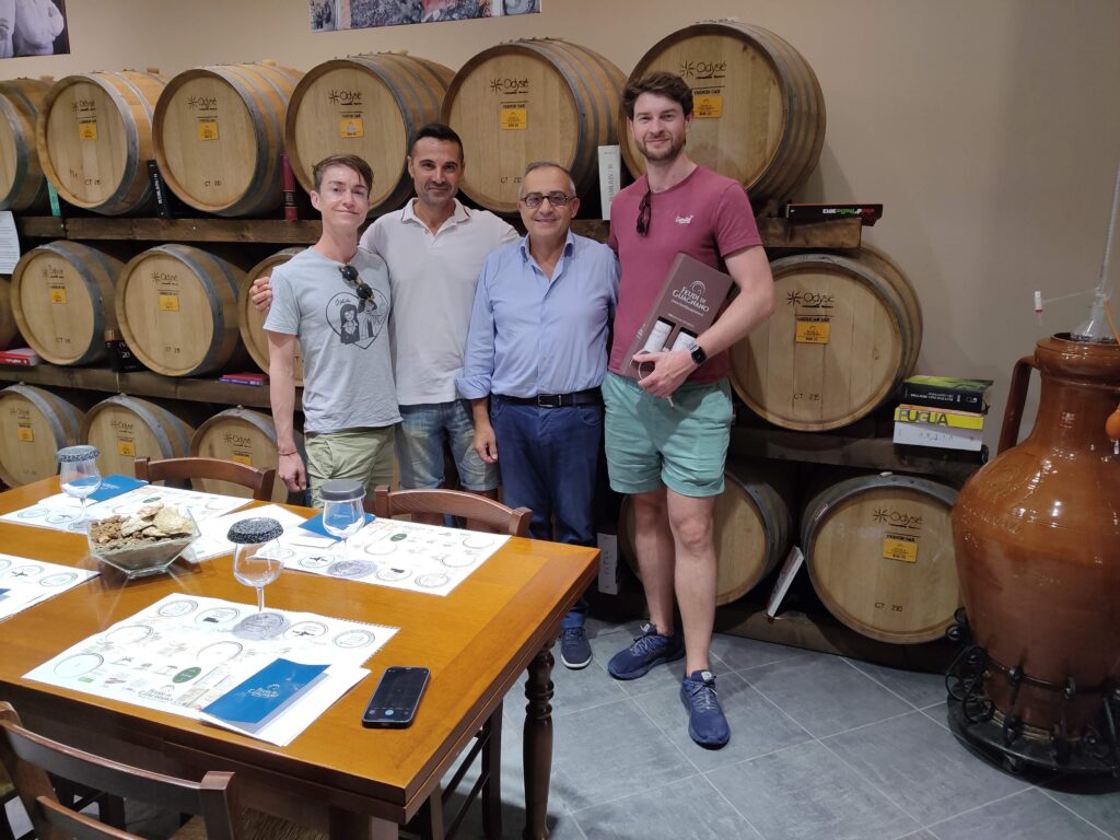 Gianvito Rizzo, owner of Feudi di Guagnano winery, posing with three satisfied international clients in the barrel cellar. They are standing in front of rows of oak aging barrels. One client holds a wine gift box. In the foreground, a tasting table is set with professional placemats, glasses, and local taralli.