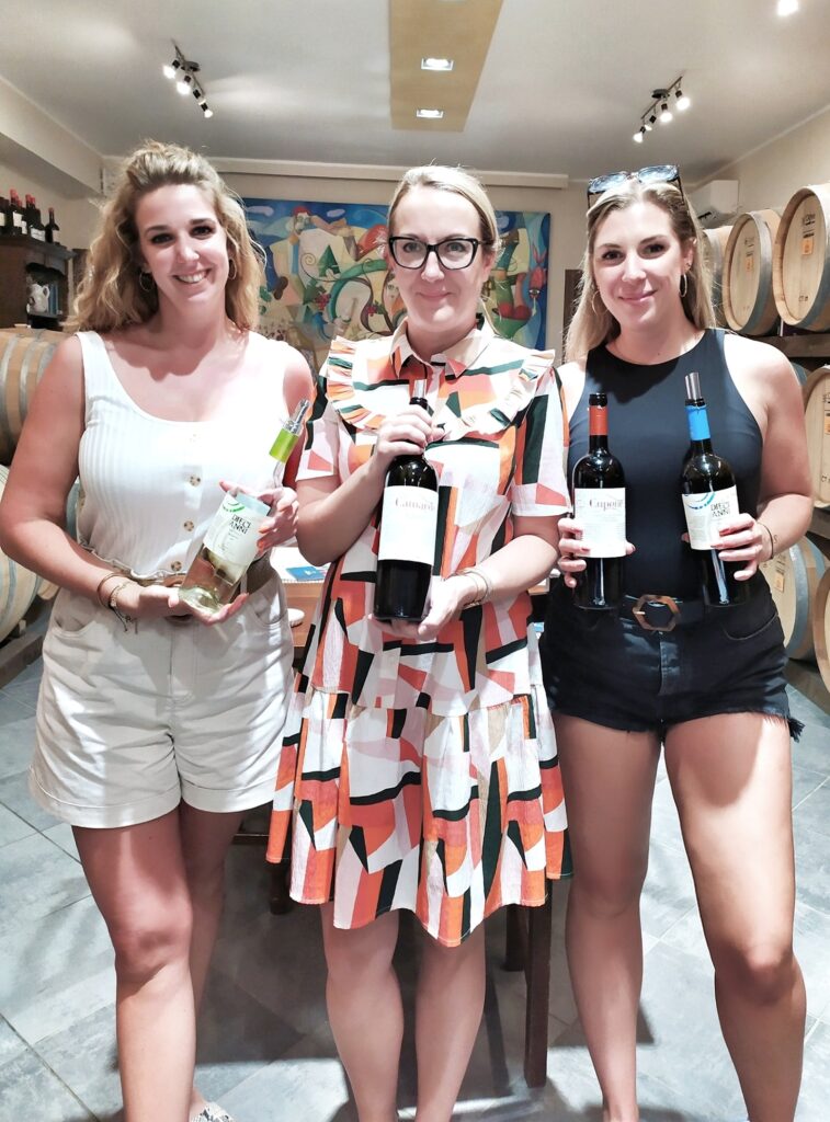 Three smiling young women posing together in the aging cellar of Feudi di Guagnano winery. They are proudly holding bottles of Salento wine, including "Diecianni" and "Cupone," with professional wine pourers attached. They stand in front of stacked oak barrels and a colorful modern mural.