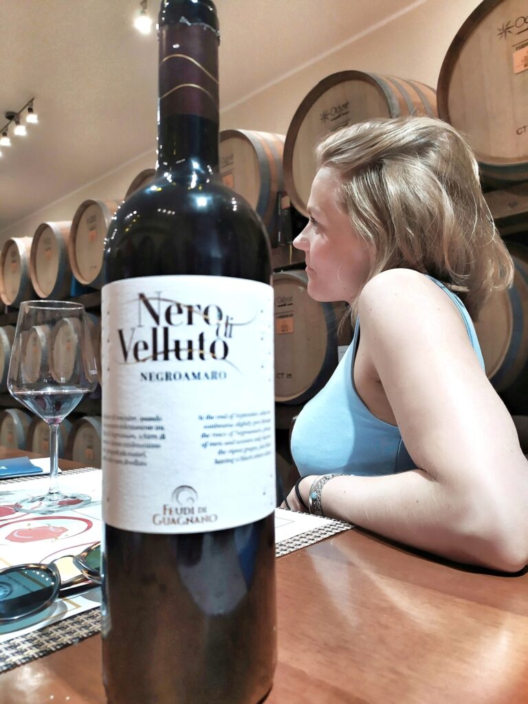 A close-up focus on a bottle of "Nero di Velluto" (Black Velvet) Negroamaro by Feudi di Guagnano. The elegant white label is prominent in the foreground, with a glass of red wine and a blonde woman looking towards the barrels in the blurred background. The setting is the aging cellar with oak barriques.