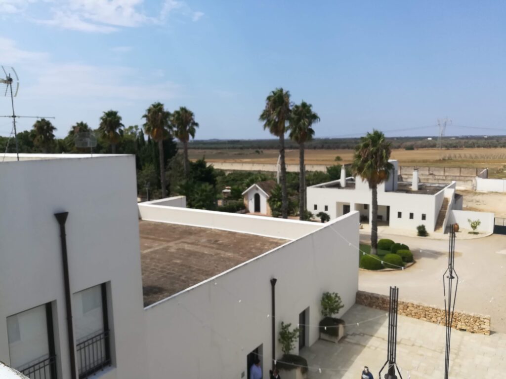 An elevated aerial photograph capturing the modern, white geometric architecture of Masseria La Cornula in Copertino, Puglia. The scene includes flat, stepped roofs, a clean paved courtyard, towering palm trees, and a small chapel. The agricultural fields of Salento stretch out in the background under a partly cloudy blue sky.