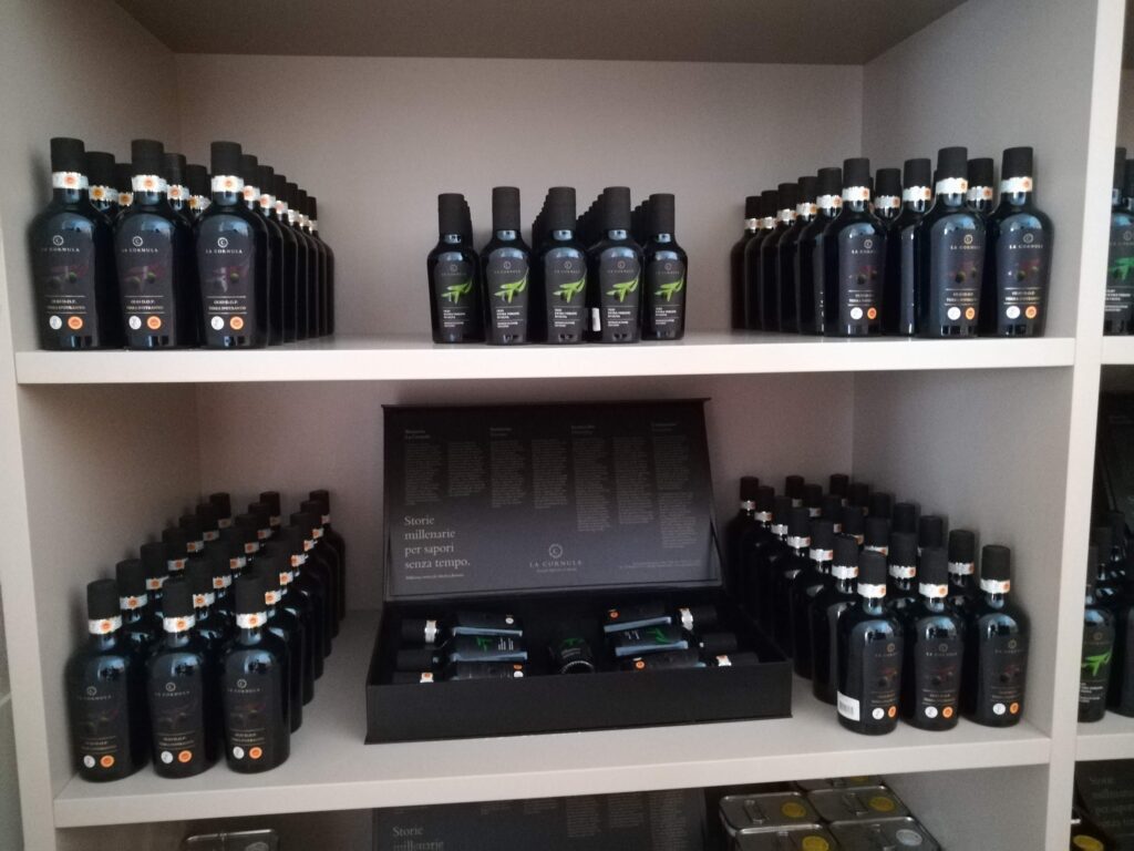 A professional indoor photograph of neatly arranged rows of elegant dark glass bottles on shelves, showcasing Masseria La Cornula's "Terra d'Otranto" extra virgin olive oil. In the center, a black luxury gift box is open, displaying smaller tasting bottles with an etched quote about millenary stories and timeless flavors. The setting is the masseria's gourmet shop.