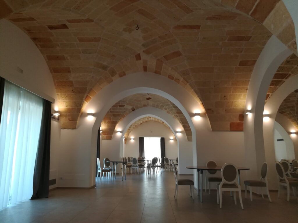 The interior of a spacious dining or event hall at Masseria La Cornula in Puglia, featuring traditional star-vaulted ceilings made of local Pietra Leccese stone. Modern tables and elegant grey chairs are arranged under the historic arches, with soft wall lighting enhancing the warm texture of the stone.