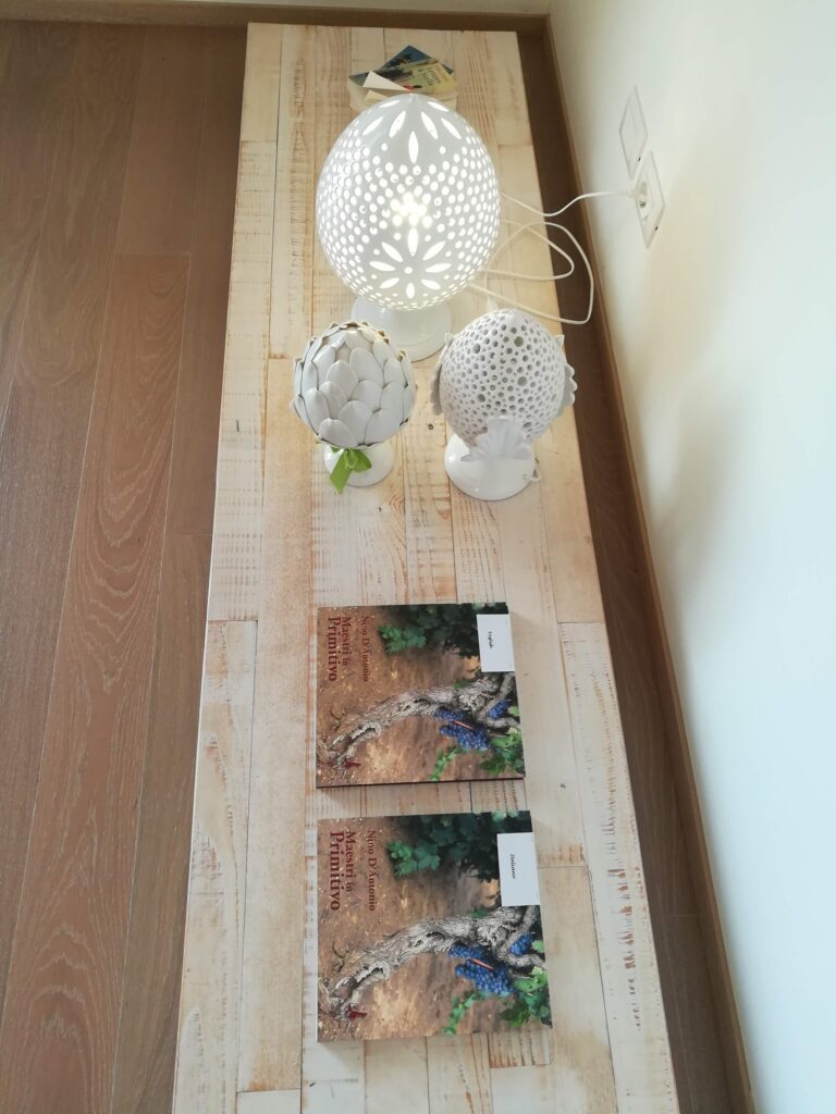 A high-angle shot of a wellness area in Masseria La Cornula, Puglia. On a light wood table, there are three white hand-painted ceramic lamps with intricate cut-out patterns, including a traditional artichoke shape. Two wine books titled "Maestri in Primitivo" are placed neatly on the table.