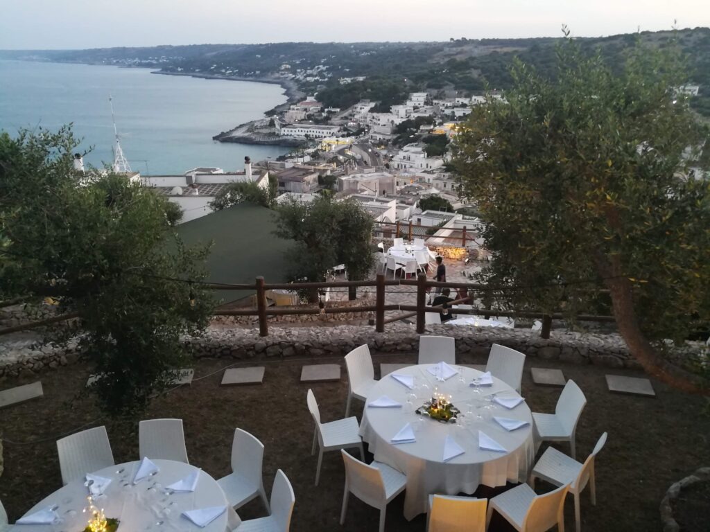 A high-angle landscape photograph capturing another sweeping, high-angle panoramic view of the wide, turquoise Castro Bay (Baia di Castro) on the Salento coast of Puglia, Italy, taken from the ancient upper castle terrace. In the foreground, numerous intricately prepared round tables with white linens, complex table settings, and a glass receiving a pour are arranged for a VIP event, framed by ancient limestone walls and Roman-era columns. A young woman with a green dress sits on the extreme edge of the weathered cliff on the left, her back to the camera, gazing out over the water. The bay is framed by extensive limestone formations, Roman and medieval archaeological ruins (Grotta della Poesia site), and dense Mediterranean maquis. Traditional white buildings line the distant shore. The sun is high, casting crisp shadows under a wide, partly cloudy blue sky, highlighting the crystal-clear water and ancient stone. The composition leads the eye from the foreground figure to the vast coastal panorama and the exclusive event setting.