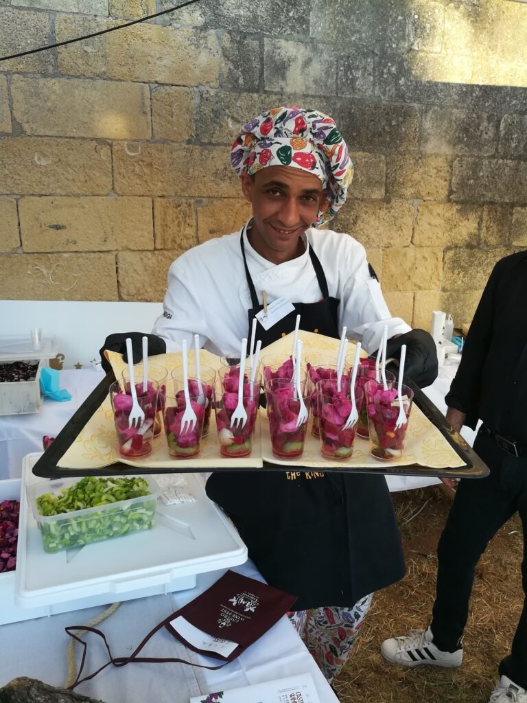A friendly chef in a colorful patterned toque and black apron smiles while holding a large tray of gourmet appetizers at the Castro Wine Fest in Salento, Puglia. The tray features individual glasses filled with a vibrant magenta-colored beet and vegetable ceviche or salad, each with a small white fork. In the foreground, a wine tasting pouch and fresh celery are visible on a white table against a rustic limestone wall.