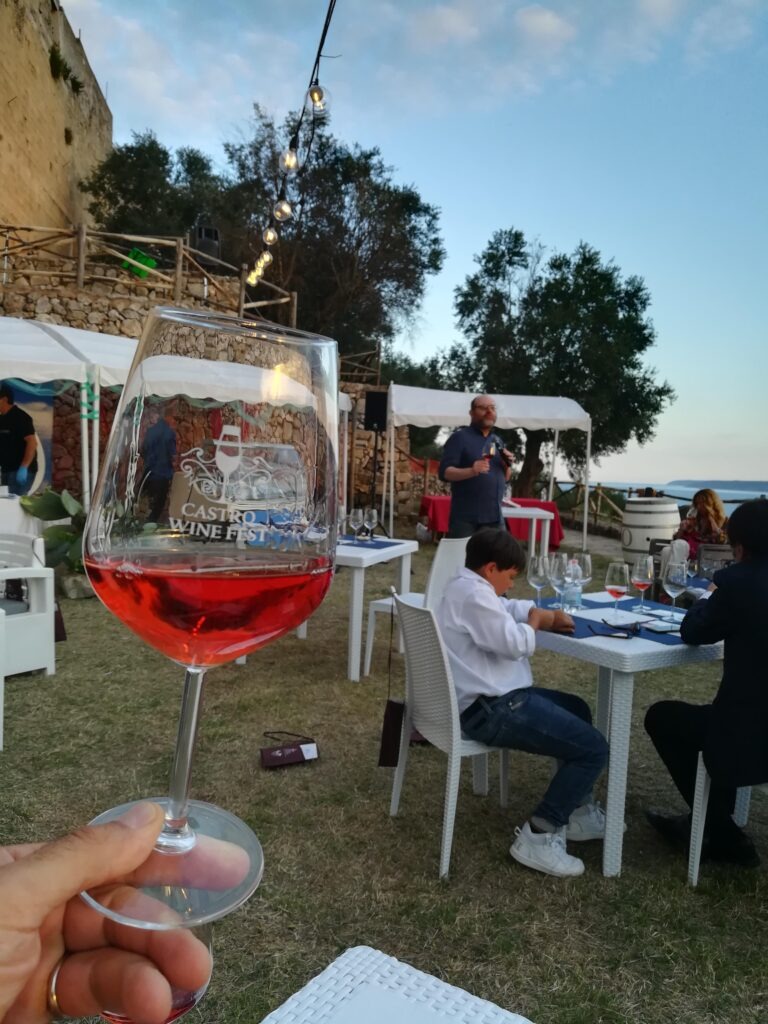 A creative close-up of a hand holding a glass of bright pink Salento Rosé wine. Through the crystal glass, the reflection shows the historic stone walls of Castro's ancient castle and the coastal landscape. In the background, a wine masterclass is in progress with participants seated at white tables on a lawn, under a twilight sky during the Castro Wine Fest.