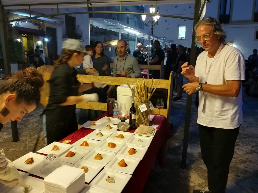 Michelin-starred chef Giorgio Locatelli (with curly grey hair, glasses, and white shirt) in a private, authentic setting during the Castro Wine Fest in Salento, Puglia, Italy, meticulously preparing a complex appetizer with a tweezer. He stands behind a professional stainless steel and marble preparation counter in a well-lit outdoor evening kitchen, surrounded by fresh ingredients (leafy greens, specific Salento red prawns, scampi, mussels, clams), diverse plating elements, and specialized tools. Behind him, the festive lights, architectural details of the ancient castle walls, other attendees, and a distant sea view are softly blurred, creating a focused, high-energy atmosphere that emphasizes Locatelli's precision and the high standard of the event.