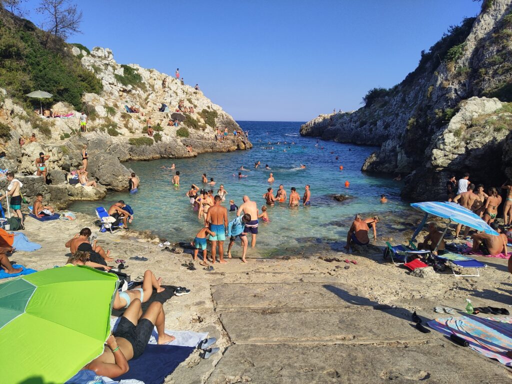 Looking out from a concrete ramp onto the narrow, rocky Acquaviva di Marittima cove (Cala dell'Acquaviva) near Diso, Salento, Puglia, Italy, featuring turquoise and deep blue water, steep, shrub-covered karst cliffs, and numerous swimmers and sunbathers under a bright, clear blue sky.