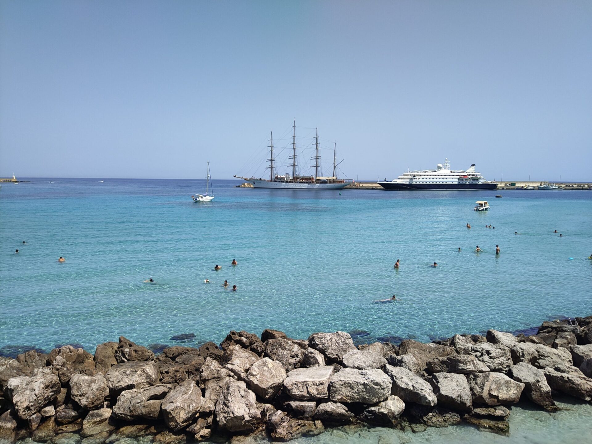 Otranto city beach with crystal clear turquoise water, harbor and cruise ships at anchor