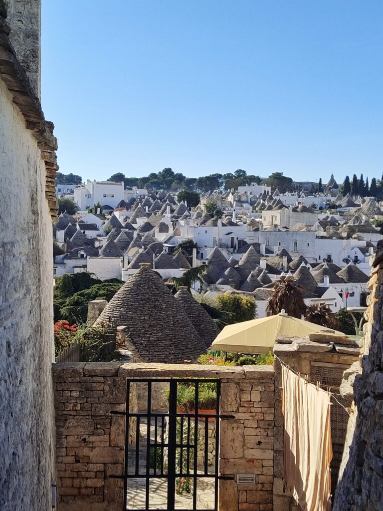 A sweeping panoramic view of the traditional stone-coned Trulli rooftops in Alberobello, Valle d'Itria, Puglia, Italy, under a clear blue sky. In the foreground, a rustic stone stairway with a black iron gate and drying laundry on a clothesline frames the historic, whitened residential quarter, emphasizing local life. A natural tree line borders the town in the distance. This unique Apulian architecture is a UNESCO World Heritage site.