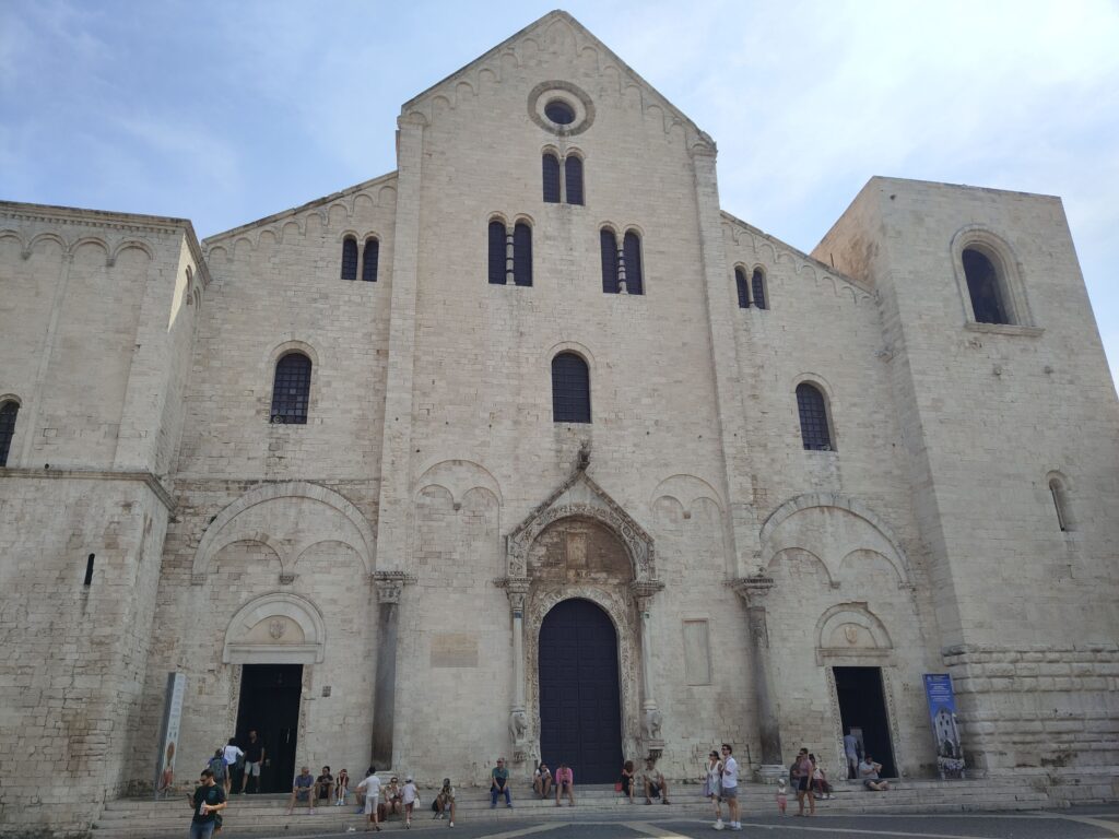 A high-definition photograph capturing the grand Romanesque facade of the Basilica di San Nicola in Bari, Italy, during a partly cloudy day. In the foreground, a detailed, ornate stone plinth is held by a hand wearing a wedding ring. On the plinth, a polished glass display box containing genuine mortal remains and a detailed text panel, alongside small, ornate reliefs of Saint Nicholas blessing children and Santa Claus on a sleigh. Directly behind, a vibrant outdoor market stall is visible, selling traditional Christmas items and 'Nicolaus' branded goods, with festive red and green ropes in front. The stone-paved square and surrounding buildings are clearly visible. The lighting is sunny and detailed, emphasizing the history, cultural blend, and craftsmanship. The view is comprehensive and inviting.