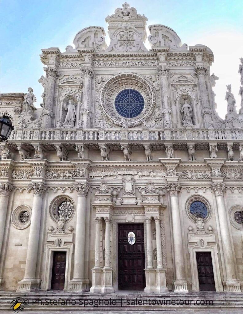 A full-frontal, detailed architectural photograph of the ornate facade of the Basilica di Santa Croce in Lecce, Puglia. The image highlights the magnificent central rose window, the complex balcony supported by zoomorphic and allegorical figures, and the intricate carvings of the "pietra leccese" limestone. The blue sky is visible in the background.