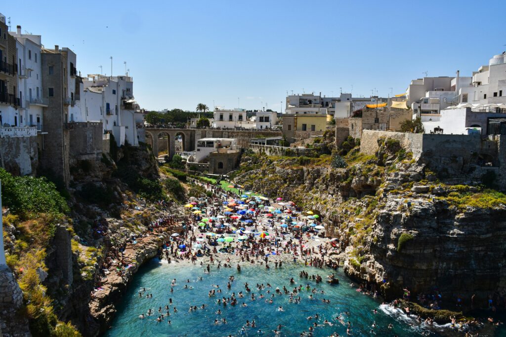 An iconic high-angle photograph capturing the stunning Lama Monachile beach (Spiaggia di Polignano a Mare) from above. The composition features naturally carved limestone cliffs that frame the narrow pebble beach, teeming with sunbathers and colorful umbrellas, and crystal-clear turquoise and deep azure Adriatic Sea water dotted with swimmers. Majestic white medieval buildings and ancient defensive walls line the extreme cliff edges, with traditional stone architecture leading to the historic center under a bright blue sky. An ancient stone bridge spanning the cove provides a dramatic backdrop. A hand holds a ring-adorned hand on a wooden railing in the foreground. The composition leads the eye from the foreground figure to the vast coastal panorama and historic borgo.