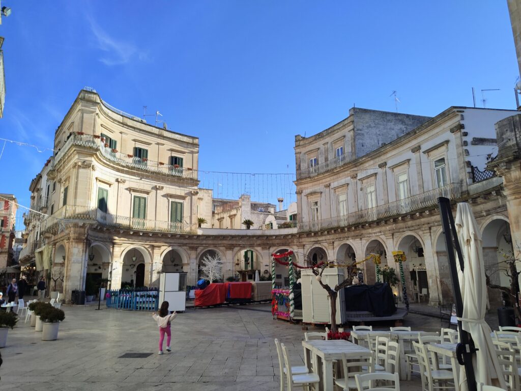 A wide-angle view of the iconic Piazza Maria Immacolata in Martina Franca, Puglia, Italy. The square is characterized by its semi-circular neoclassical buildings with elegant porticos, balconies with green shutters, and outdoor cafe seating with white wooden tables. A child in a pink outfit is seen in the center of the stone-paved square under a clear blue sky.