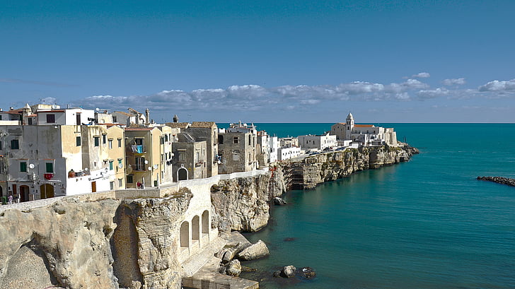A panoramic wide shot of the historic white city of Vieste, built directly on the rugged limestone cliffs overlooking the deep turquoise Adriatic Sea under a clear blue sky. The Chianca Amara and the iconic San Francesco Church are visible at the tip of the peninsula.