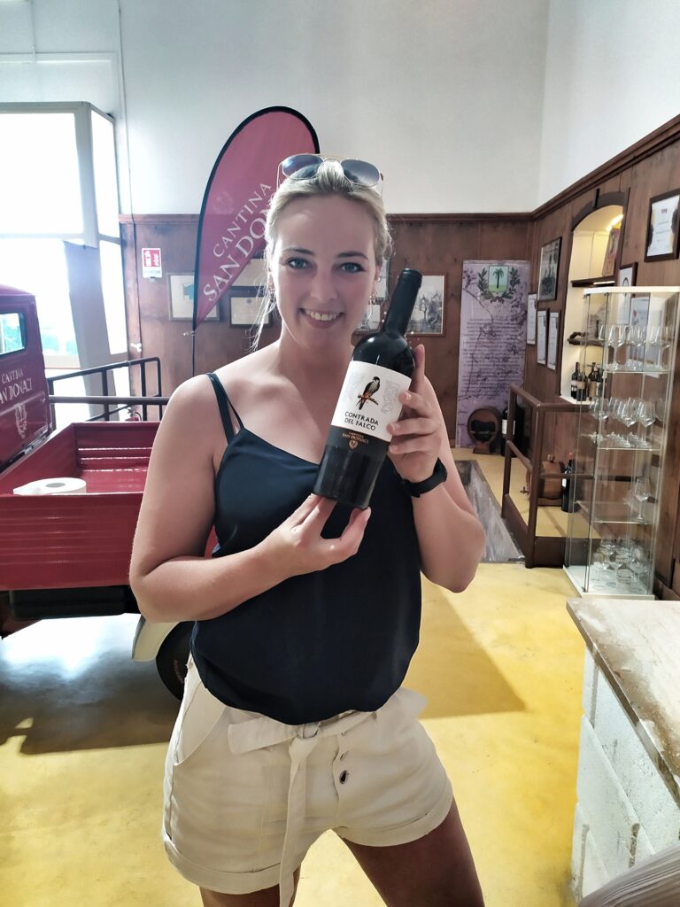 A smiling young woman holding a bottle of "Contrada del Falco," a premium Negroamaro, Primitivo, and Malvasia Nera red wine blend, in the charming, history-filled wine shop of Cantina San Donaci in Salento, Puglia, Italy, featuring a red three-wheeler and a historic stone press pit.