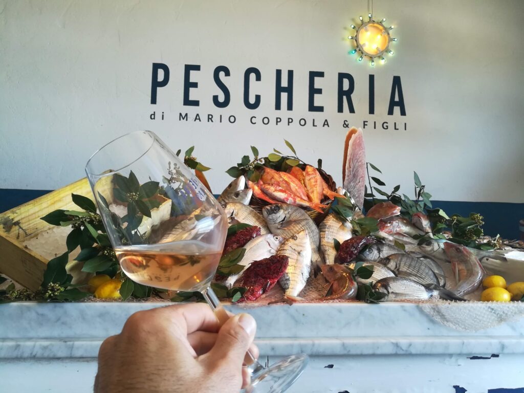A high-angle view of a dramatic, large fresh fish display on a marble slab in 'La Pescheria di Mario Coppola & Figli' restaurant in Tricase Porto, Puglia, Italy. The display features a hand holding a glass of amber-colored Rosé wine in the foreground, with a large assortment of local Mediterranean wild-caught fish, lemons, and bay leaves on a fishing net. The restaurant name and logo are on the white wall, with a specific decorative neon light above.