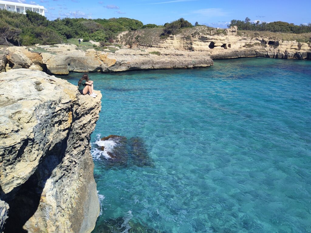 A sweeping, high-angle landscape photograph looking across the wide, circular bay of Roca Vecchia, near Otranto, Salento, Puglia, Italy. The image features incredibly clear, multi-faceted turquoise and aqua-blue water under a vast, partly cloudy blue sky. On the left, a solitary young woman with a green t-shirt sits at the extreme precipice of a high, rugged calcarenite limestone cliff. In the distance, the extensive calcarenite rock formations with natural sea caves, arches, and scattered archaeological ruins are visible, cradling the bay. Dense Mediterranean maquis and traditional white buildings line the distant shore.