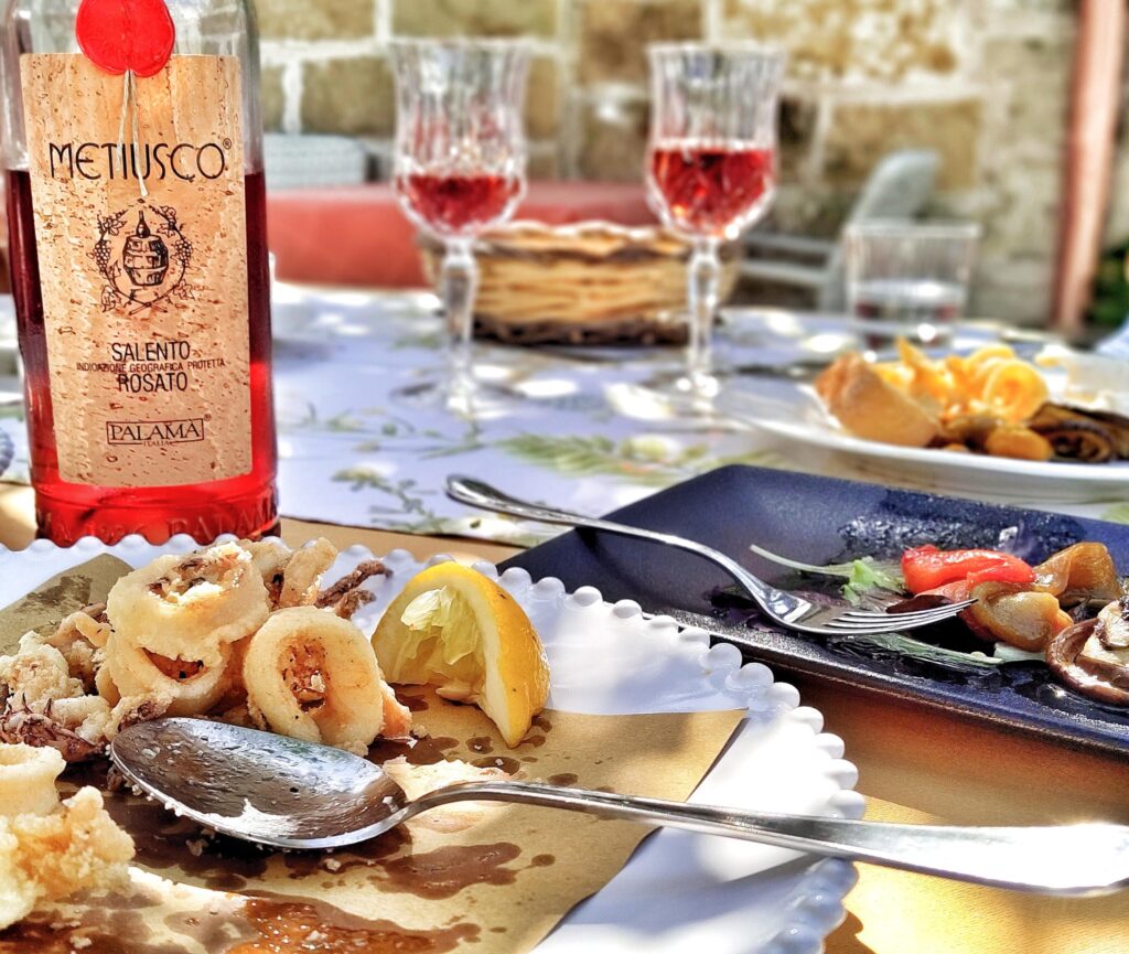 A tranquil, private dining table in the beautiful inner garden of Ristorante Da Amedeo in Castro, Salento, featuring a bottle of Metiusco Salento Rosato wine, two glasses of rosé, and a plate of fresh fried calamari under a natural canopy.