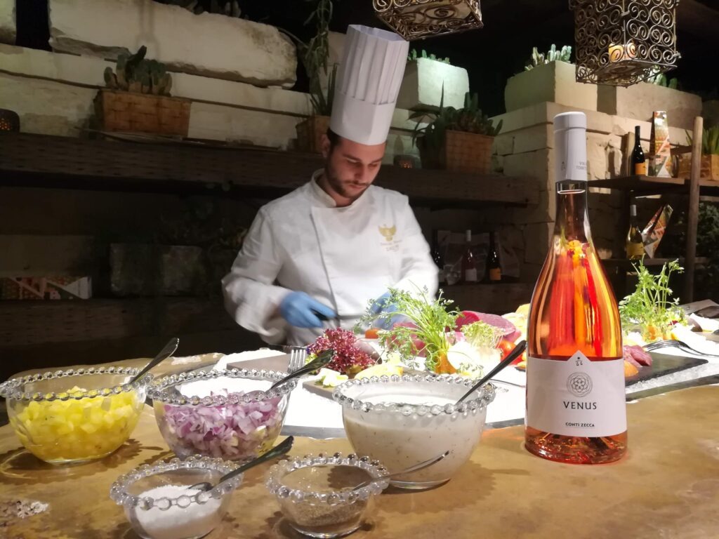 A professional chef with a tall white hat and gloves in a rustic, well-lit kitchen in Salento, Puglia, Italy, meticulously preparing an appetizer (antipasto) using fresh, local Mediterranean ingredients. In the foreground, bowls of chopped pineapple, red onion, a white sauce, and spices are visible, next to a full bottle of Venus Conti Zecca Salento Rosato wine. The background features ancient stone walls, more wine bottles, and potted plants.