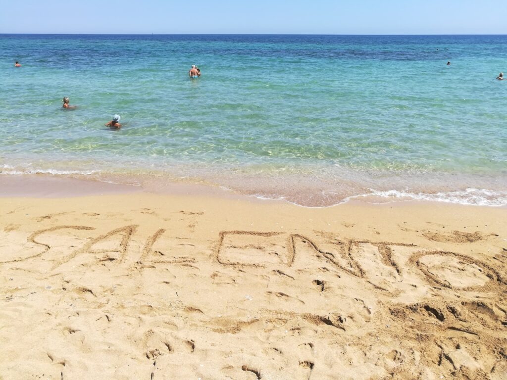 Ground-level view of the vast, golden sandy beach of San Cataldo (Spiaggia di San Cataldo) near Lecce, Salento, Puglia, Italy, featuring transparent, shallow turquoise and azure Ionian waters, people swimming in the distance, and a bright, clear blue sky. The foreground sand is marked with footprints and the large hand-written letters 'SALENTO'.