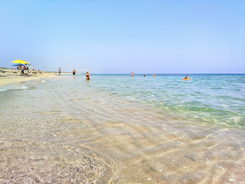 Ground-level view of the vast, white sandy beach of Alimini (Spiaggia delle Alimini) in Otranto, Salento, Puglia, Italy, featuring transparent, shallow turquoise Ionian waters, people swimming and sunbathers, and a bright, partly hazy blue sky.