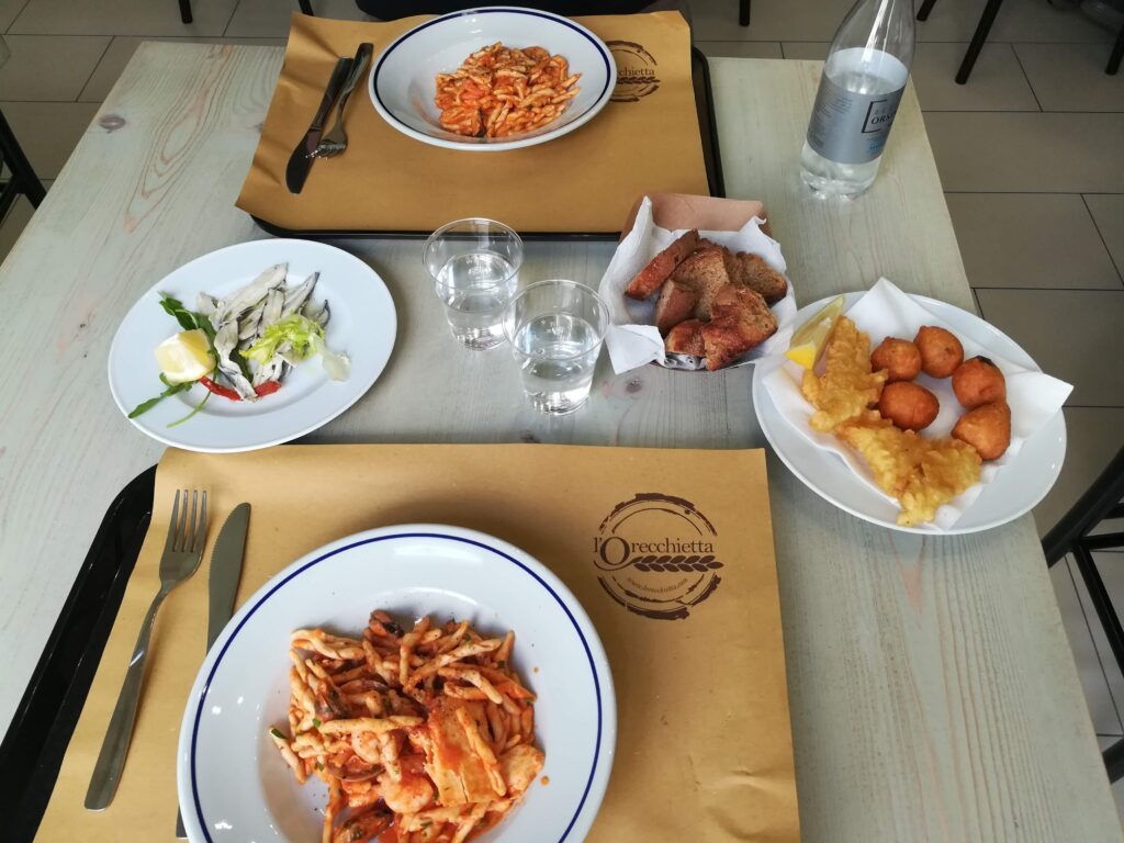 A laid table at L'Orecchietta restaurant in Guagnano, Salento, featuring two plates of handmade pasta with seafood tomato sauce, appetizers with marinated anchovies, fried pettole, and local bread on brown paper placemats.