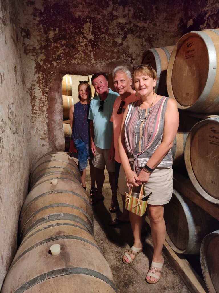 A group of four happy tourists standing in a long row inside the historic, 4-meter deep underground concrete wine fermentation tanks at Cantina San Donaci in Puglia, Italy, now converted into a barrel room for aging premium wines.