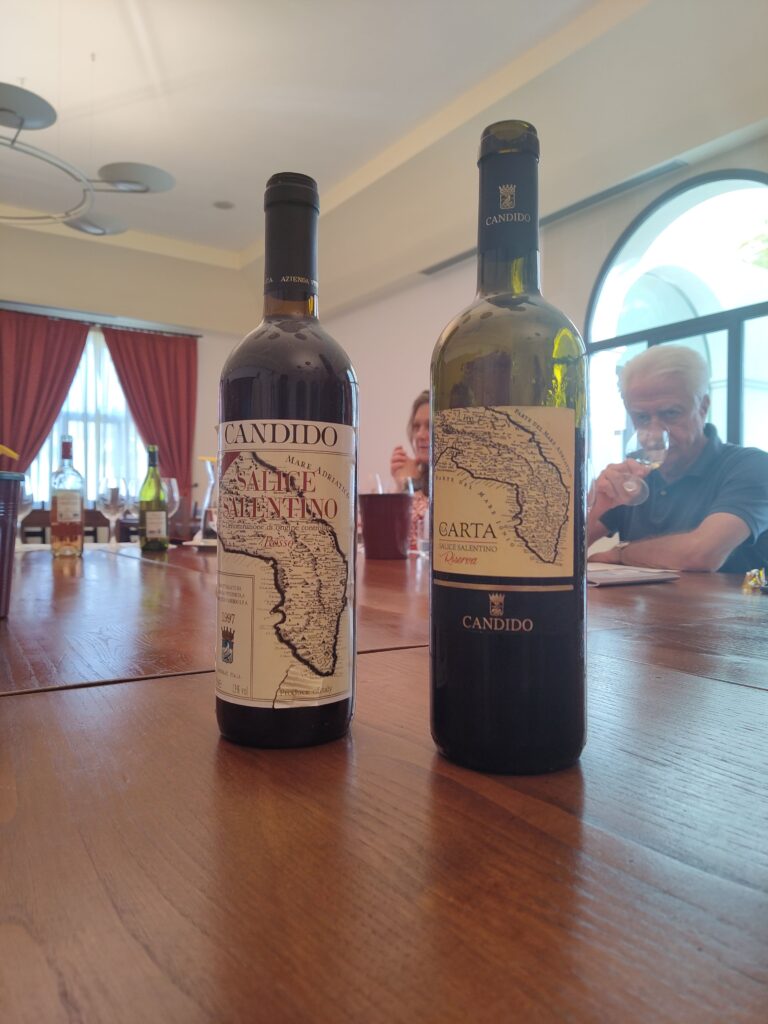 Two bottles of Candido wine on a wooden table, featuring "La Carta" Salice Salentino Riserva DOC, with winery owner Francesco Candido tasting wine in the background.