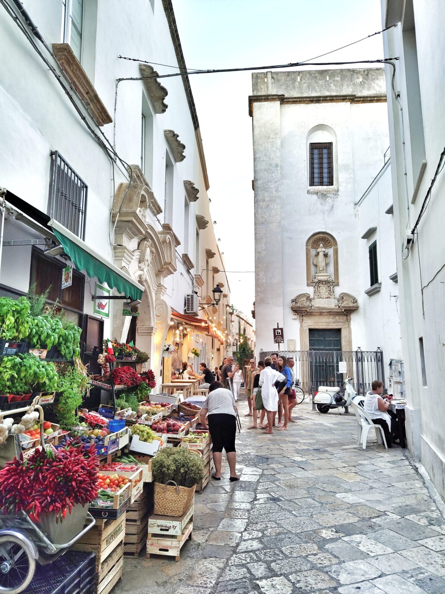 Narrow street in Gallipoli's old town with fresh fruit market and Baroque buildings in golden carparo stone